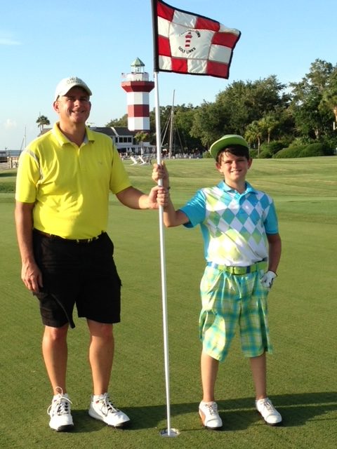 a father and son pose by a flag in front of the lighthouse on the Harbour Town Golf Links course at Hilton Head which was just named in the 10 most expensive places to retire