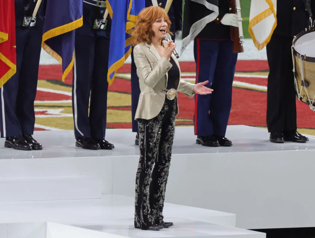 Reba McEntire Songs - Reba is singing the National Anthem at the Super Bowl wearing a tan jacket and black pants. 