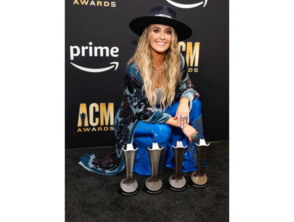 Lainey Wilson's 2024 ACM Nominations—Lainey poses with her four ACM Awards, wearing a blue and black outfit with a black hat, after the show in 2023. 